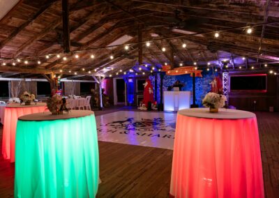 Decorative party scene: round tables with glowing green and pink skirts, string lights, and a rustic wooden ceiling; a DJ booth and festive decor in the background.