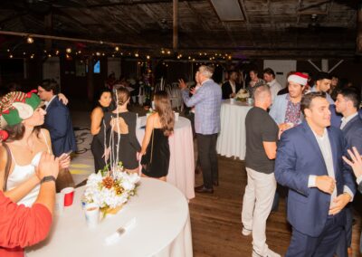 Group of guests at a Christmas party around round tables under string lights in a rustic venue.",