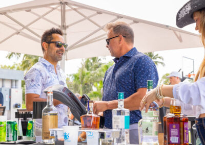 Two men in patterned shirts chat at an outdoor bar under a large white canopy, with bottles and glasses on the counter nearby.